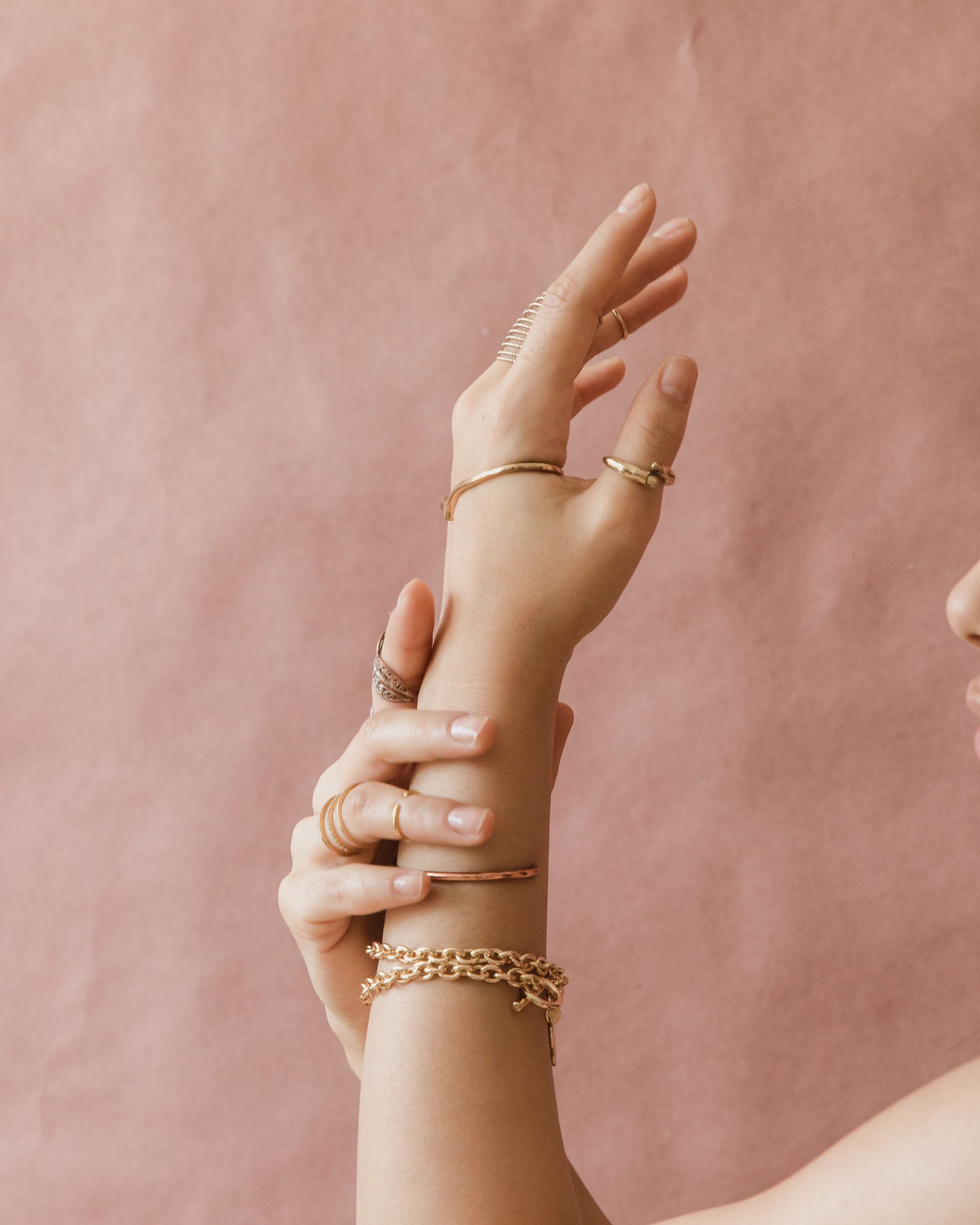 A hand wearing gold rings and a chain bracelet against a pink background.