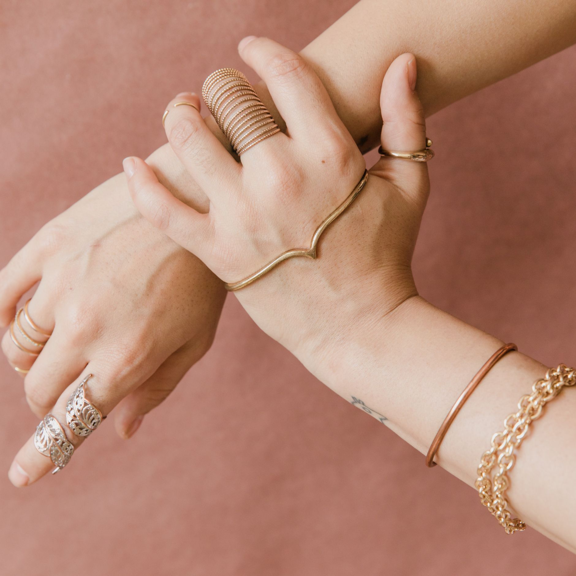 Hands wearing stacked gold rings and bracelets against a soft pink background.
